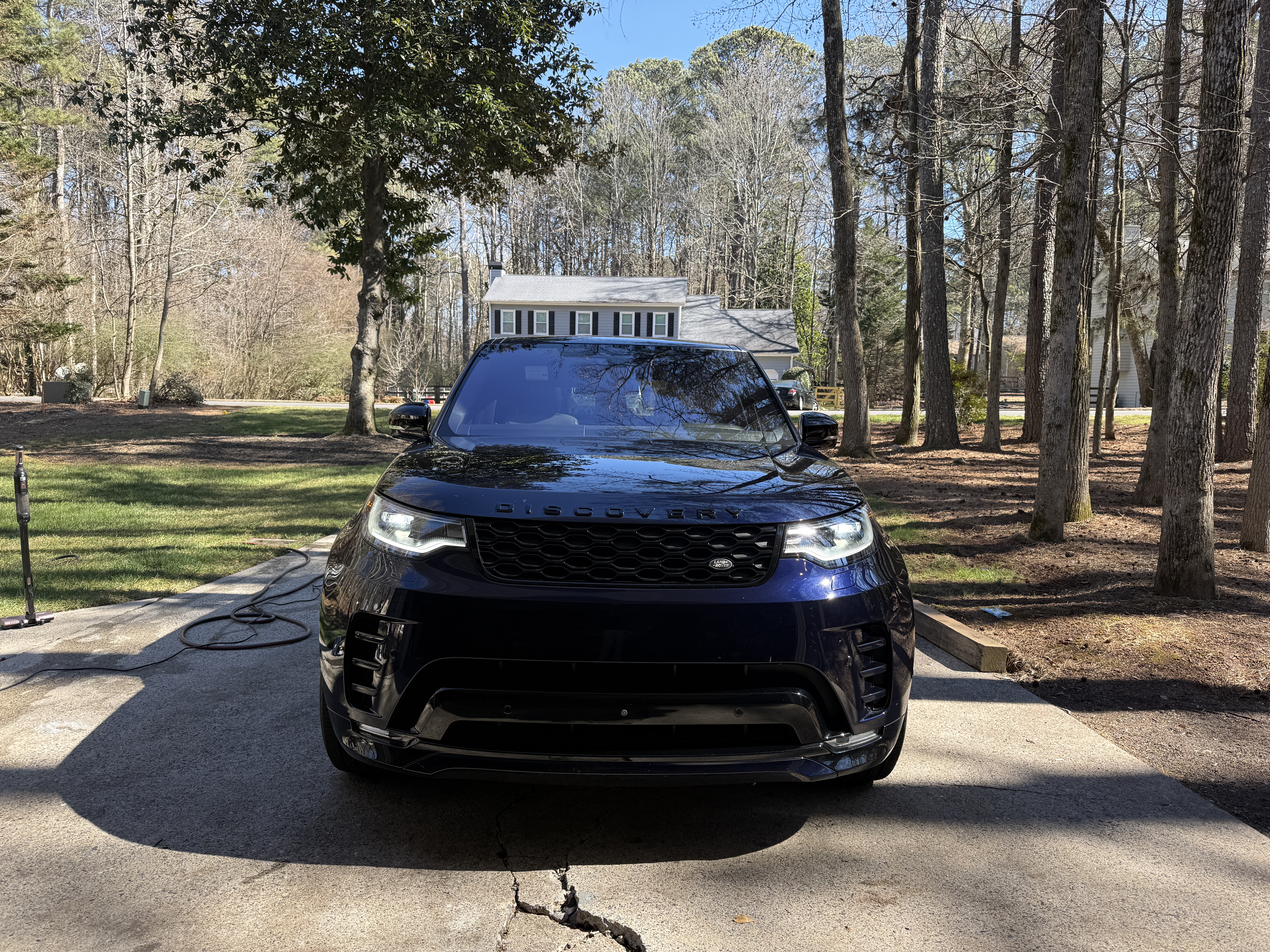 Dark blue SUV parked facing the camera on a concrete driveway after detailing; glossy hood and headlights reflecting nearby trees and sky. A coiled garden hose is to the left, a two story house and tall pine trees are in the background under clear daylight. Tone clean and professional.