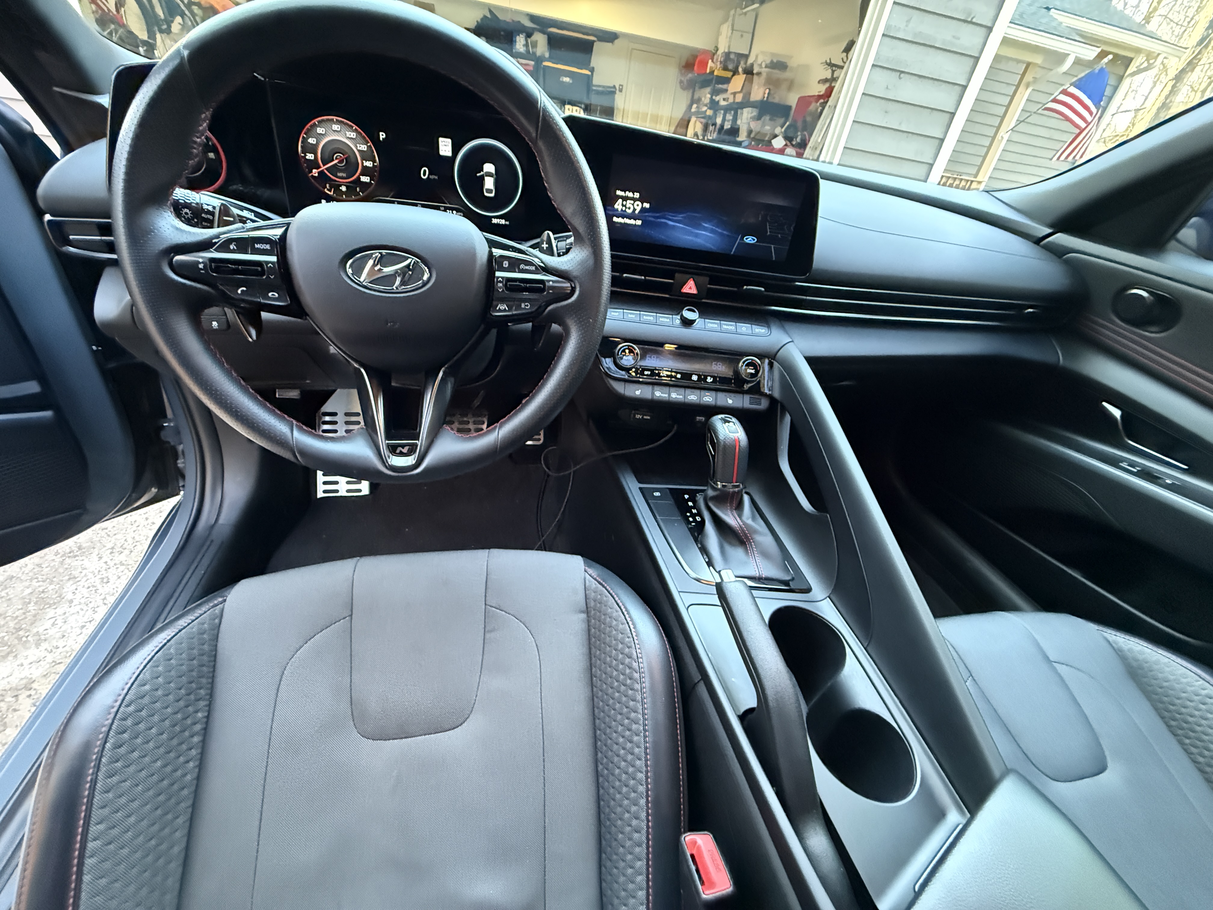 Hyundai leather steering wheel centered in the driver's cockpit with Hyundai logo on the wheel; digital instrument cluster and wide infotainment screen on the dash above the center console; automatic gear shifter, handbrake, and two cup holders in the console, plus driver and passenger seats with patterned upholstery; garage and daylight visible through the windshield; clean, polished, professional detailing result