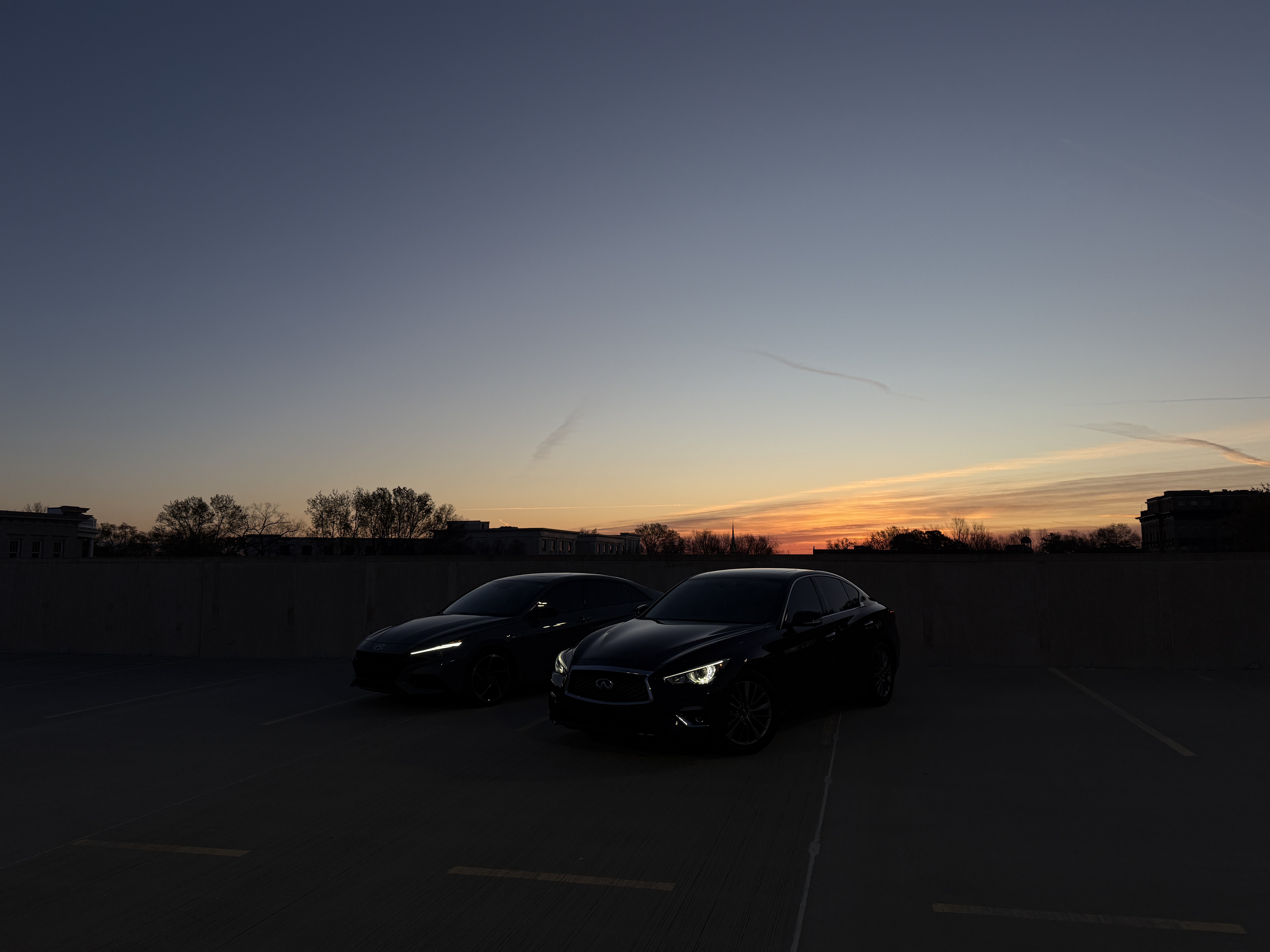Four parked cars lined side by side at night with headlights on; vehicles are stationary beneath a single streetlamp next to a multistory building with lit windows and bare trees. Wet asphalt reflects the lights. No visible text. Mood moody, cinematic, quiet, and reflective.